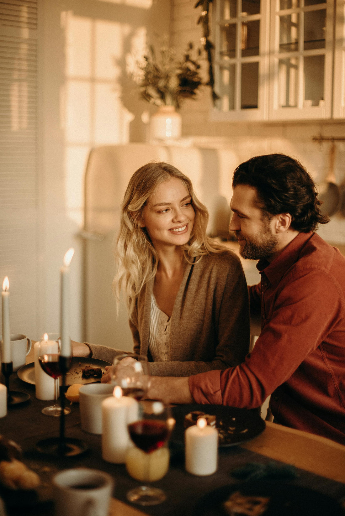 A blonde woman in a tan cardigan and white tank is smiling at a man with dark slick backed hair and a rust colored turtleneck.  They are lighting a row of candles in the center of the dining table.