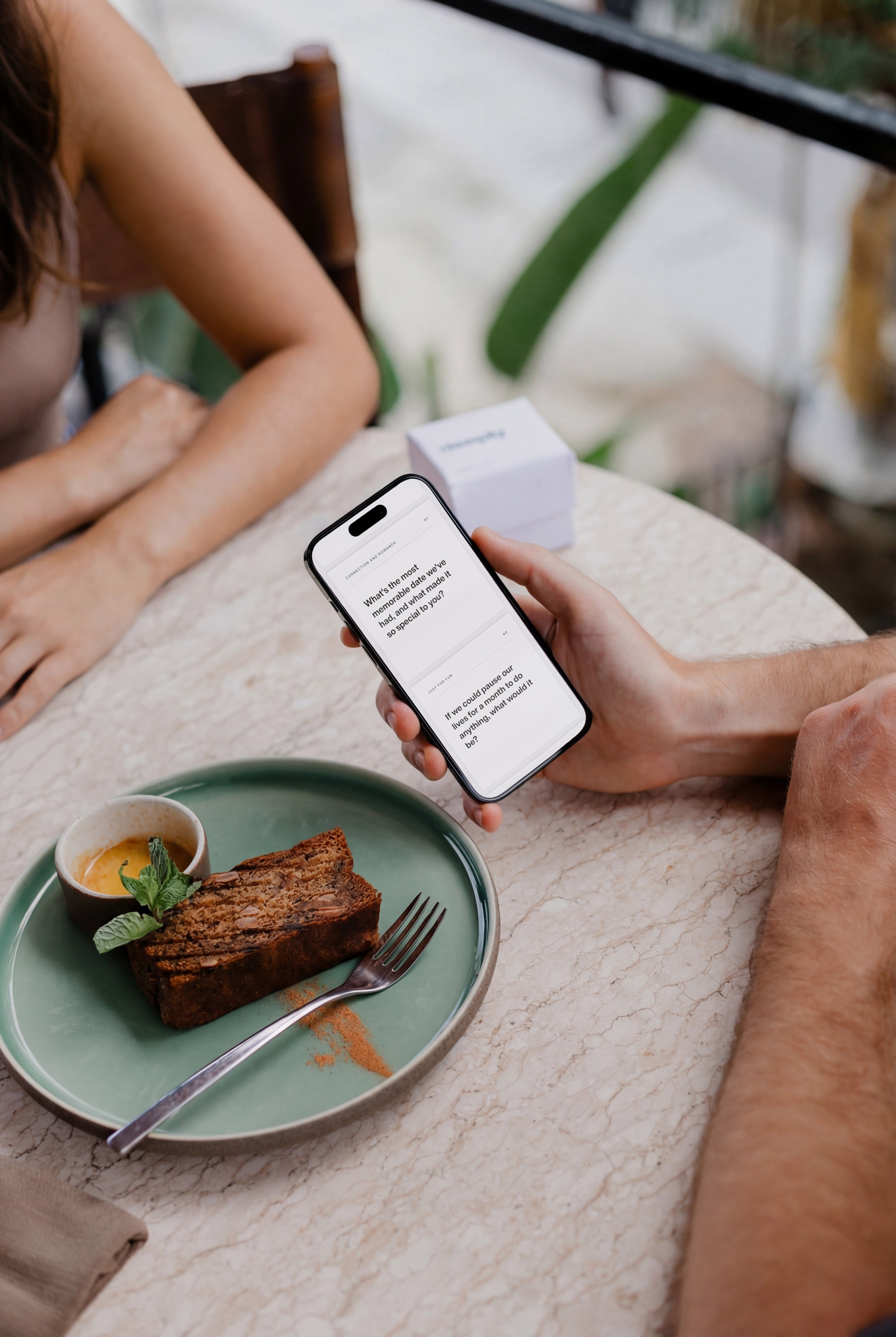 Boyfriend holding a smartphone with couples edition cards on a coffee date with a cake